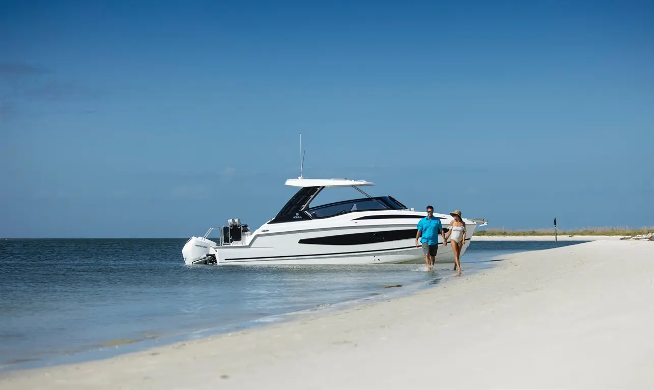 A couple walks hand-in-hand along a sandy beach, with a sleek white boat anchored nearby under a clear blue sky.